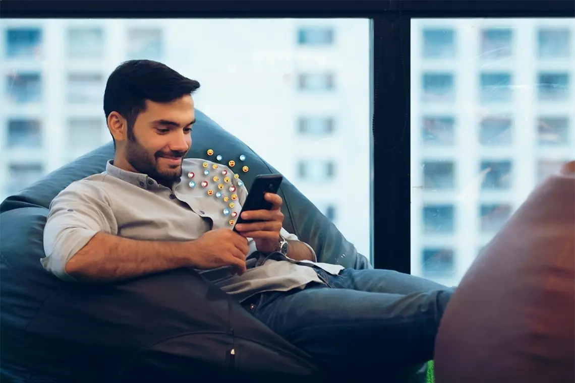 El joven profesional, sonriente, disfruta del éxito de las estrategias de marketing en su smartphone. Emojis de corazones, likes y caritas felices aparecen en el fotomontaje.