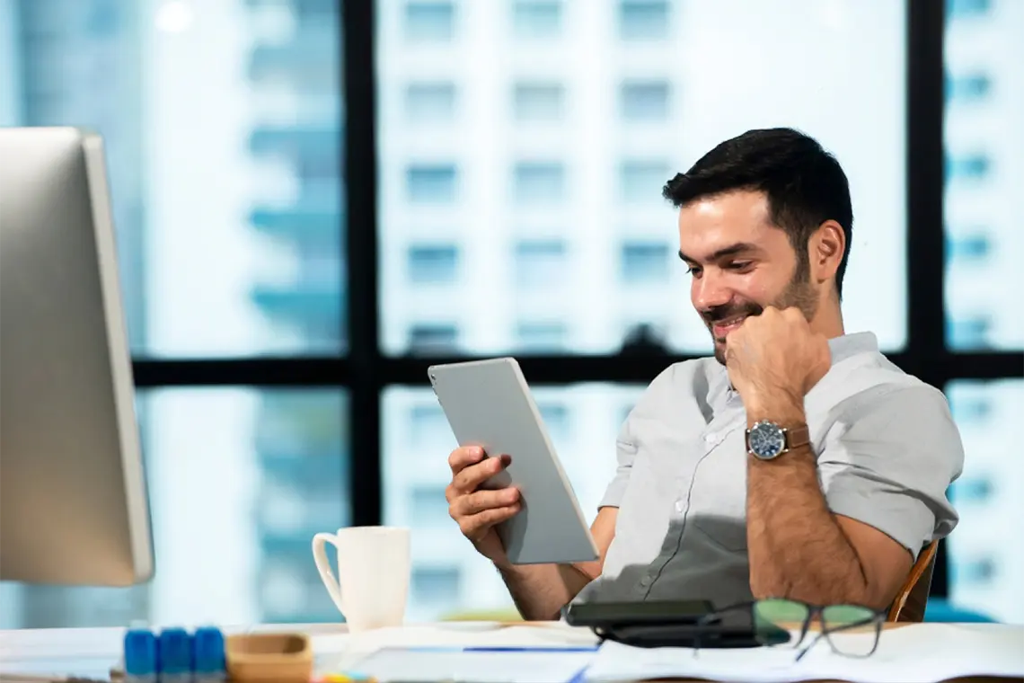 Un joven profesional, sentado frente a una PC, analiza datos de marketing en una tablet.