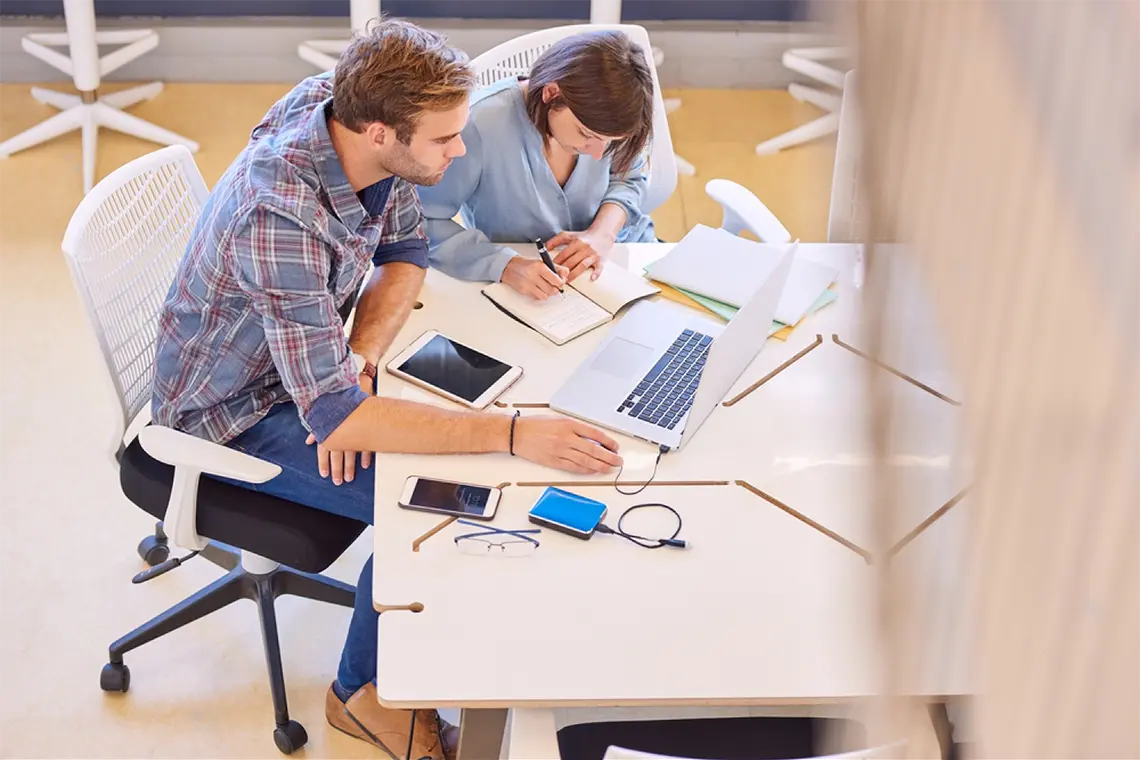 Optimización de enlaces internos Una toma más amplia del ambiente de trabajo, mostrando la laptop, tablet, cargador de móvil y celular, mientras el marketer y la supervisora discuten la optimización de los enlaces internos.