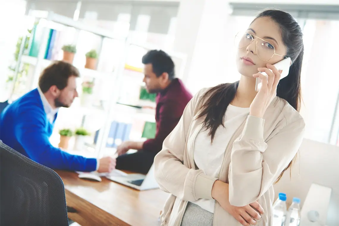 La mujer del equipo, con el celular en la oreja y mirando a la cámara, conversa con un cliente mientras sus compañeros conversan en segundo plano. [Impacto de metaetiquetas en SEO]
