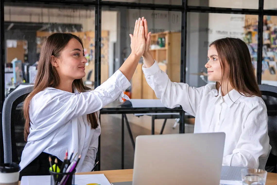 Conclusión Las dos mujeres SEO, sonrientes y con una mirada amigable, se chocan las manos en señal de haber concluido la buena gestión, de haber podido ayudar a crecer los negocios en el mercado digital.