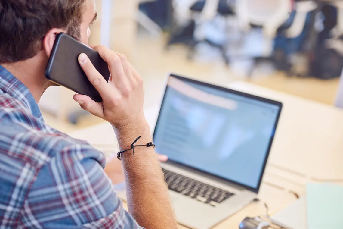 Preguntas Frecuentes El marketer sigue viendo su laptop buscando información en internet sobre la utilización efectiva de enlaces internos, atendiendo por celular las preguntas frecuentes.