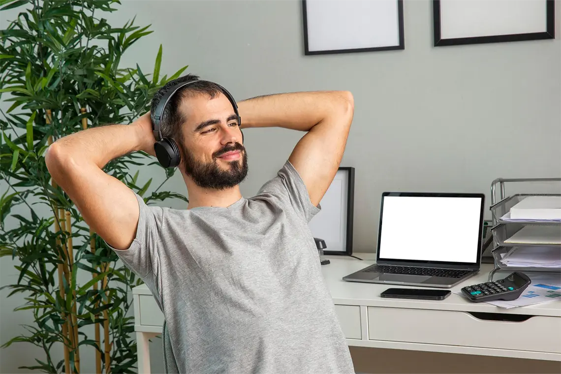Preguntas Frecuentes El hombre con las manos en la nuca, escuchando música con auriculares mientras mira de reojo su laptop que está a su costado.